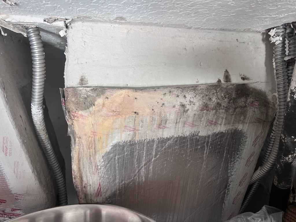 Wide view of an HVAC plenum insulation panel with dark mold growth along the top seam where the panel meets the ceiling framing