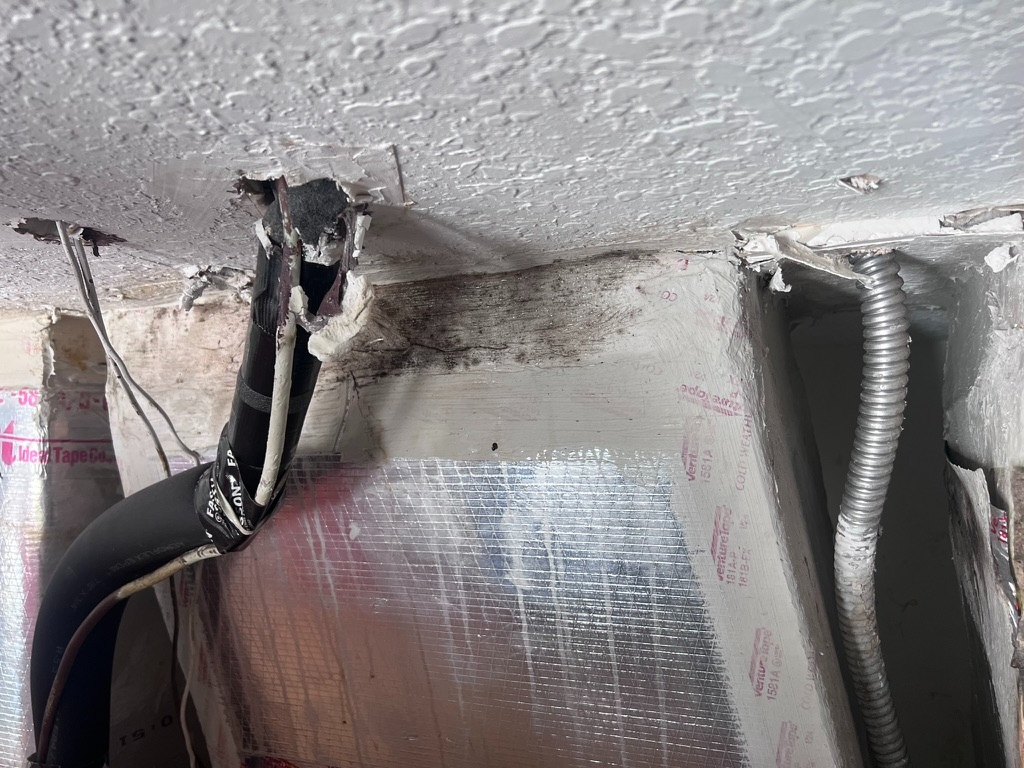 Close-up of deteriorated black HVAC sealing tape at a duct connection, with dark mold staining visible on the surrounding insulation panel